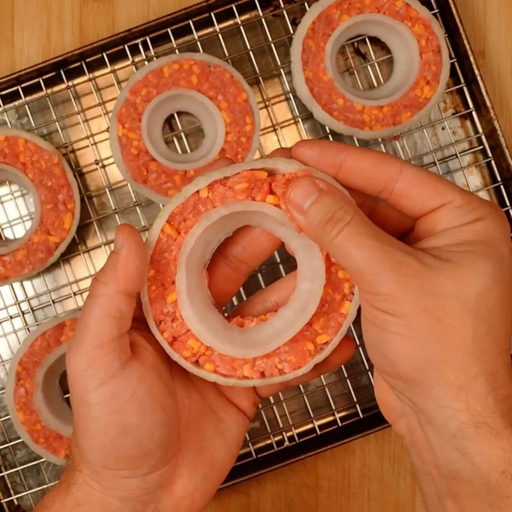 Hands holding a raw ring of ground beef with shredded cheddar packed inside a white onion ring, with more rings on a wire rack and baking sheet behind it.