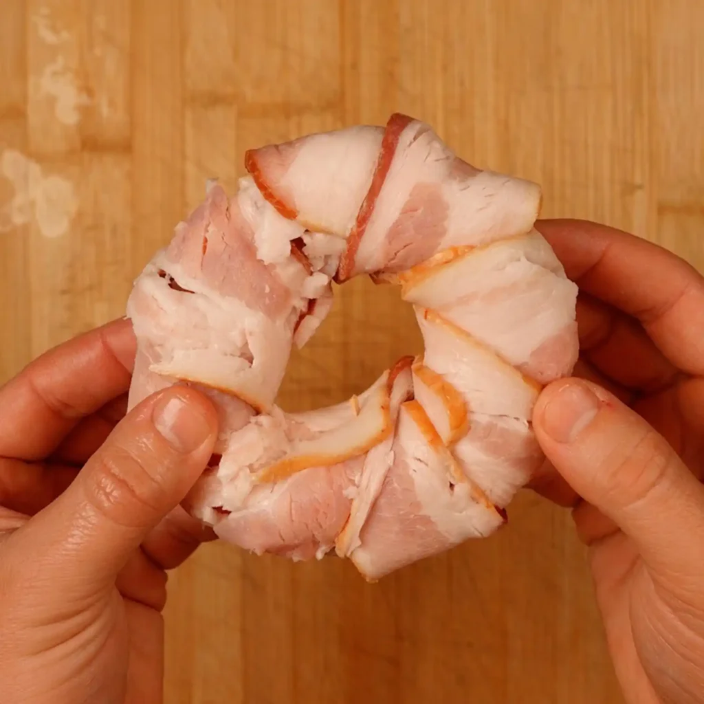 Two hands holding a ring formed from overlapping raw bacon slices over a wooden cutting board.