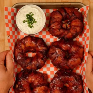 Overhead view of a tray of six glazed bacon-wrapped rings on red and white checkered paper, with a bowl of ranch dip topped with chopped green onions.