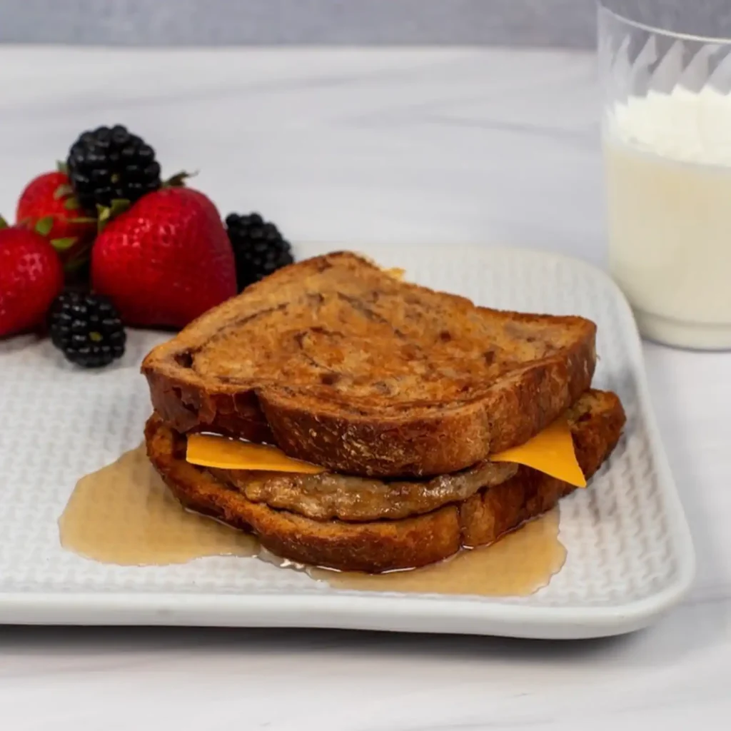 Sausage patty and melted cheddar cheese between two slices of cinnamon swirl French toast, with syrup pooling on a white square plate, served with strawberries, blackberries, and a glass of milk.